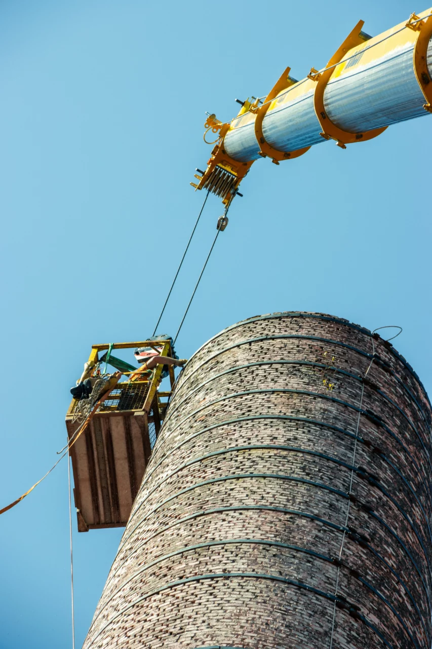 Men working in heights in a basket alongside a big factory pipe.