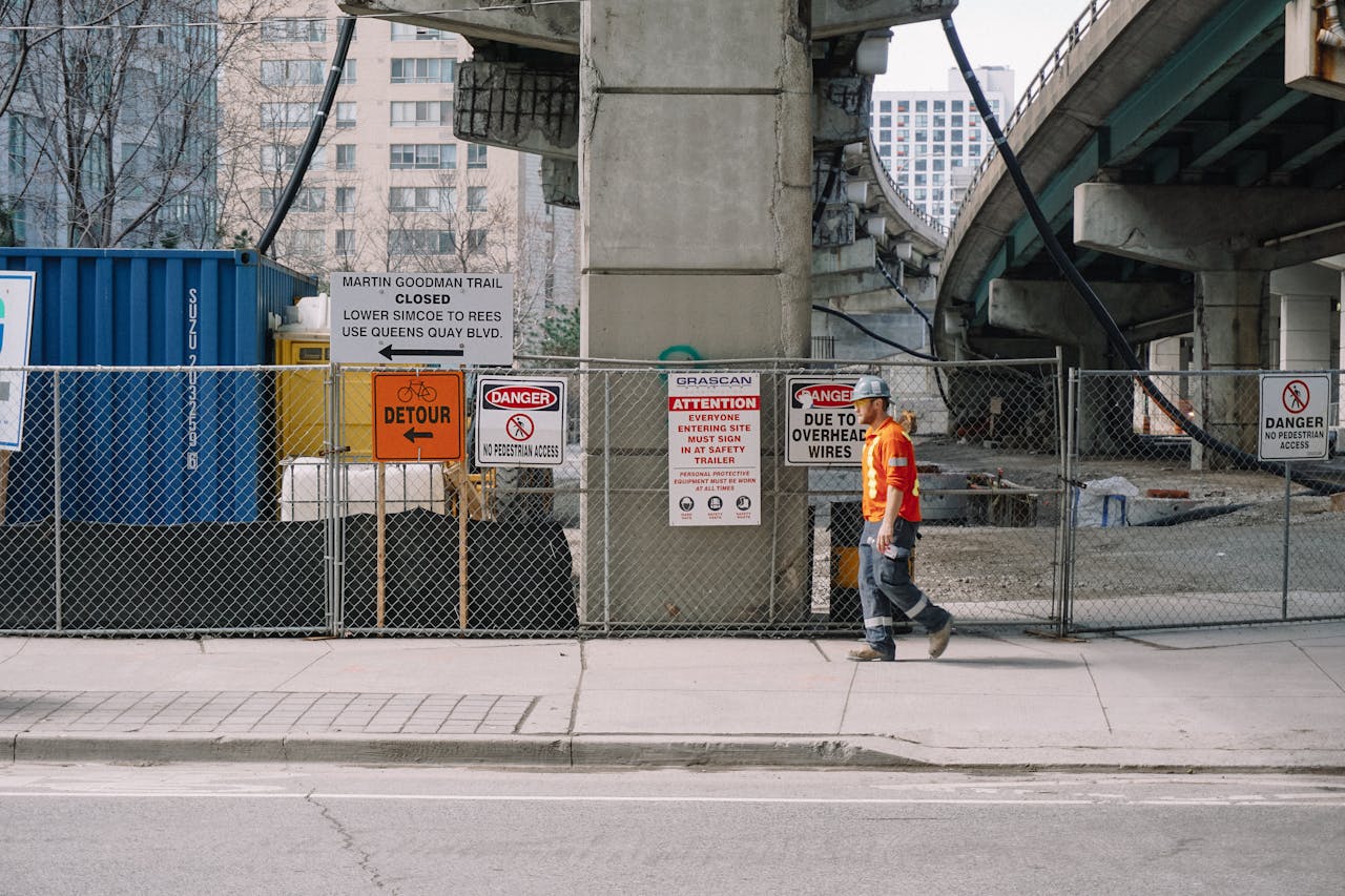 A construction worker is walking along the sidewalk, signs warning about dangers visible on the fence.