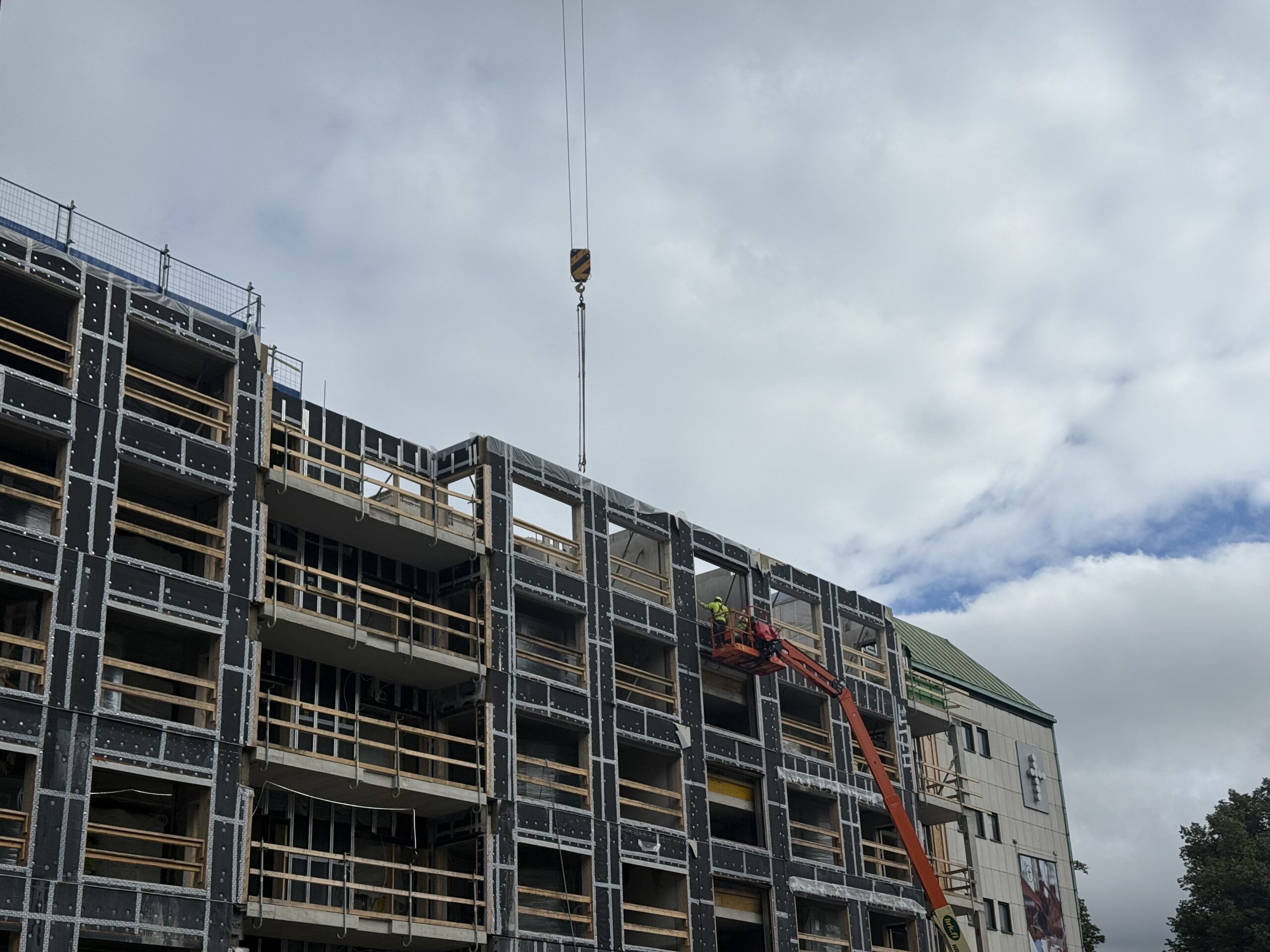 Construction workers on a cherry picker installing facade elements on a multi-story building under construction with crane on site, safety equipment visible.